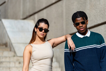 Two young models wearing stylish sunglasses pose against a concrete wall, showcasing urban fashion trends