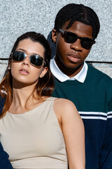 Young, stylish couple wearing sunglasses, posing next to a sleek marble wall, embodying modern urban fashion and trendy summer vibes