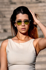 Young woman posing outdoors with sunglasses, casually touching her long hair while exuding a trendy and stylish vibe in the sunlight