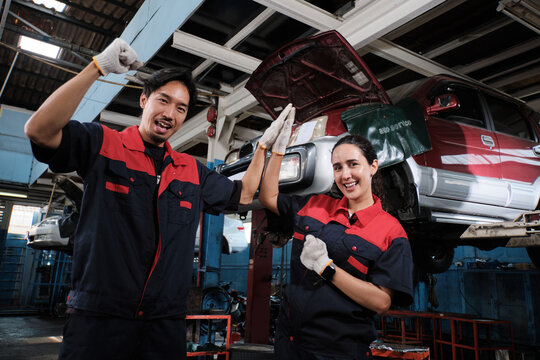 Two diverse mechanic coworkers, Latino and Asian engineer partners, were cheerful high five, car check and maintenance service at fixed auto garage, a professional transportation industry business. - Powered by Adobe