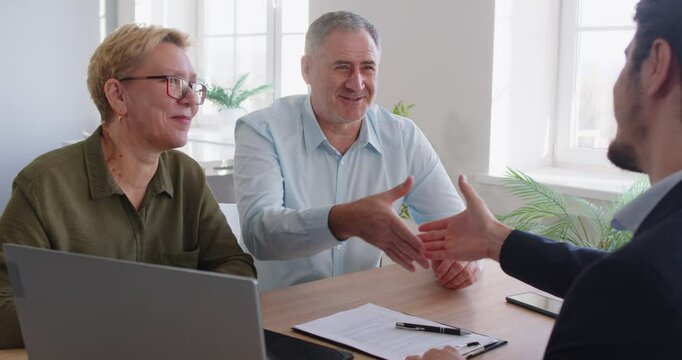 Happy smiling senior couple sitting at desk in office having consultation with male financial advisor about health insurance and shaking hands making deal. Investment in retirement concept. 4k video.