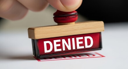 A hand holding a red rubber stamp with the word 'DENIED' on a white paper. The stamp has a wooden handle and is used for marking documents.