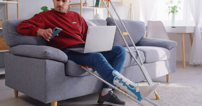 Injured man with leg in plaster cast sitting on sofa with crutches nearby, talking on smartphone and working on laptop. Young man engaged in remote work during recovery from foot trauma at home.