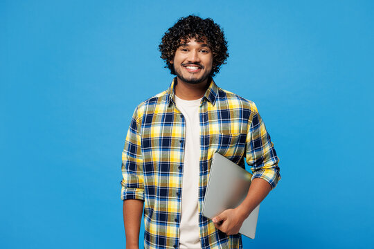 Young smiling happy IT Indian man he wears yellow checkered shirt casual clothes hold closed laptop pc computer look camera isolated on plain blue cyan background studio portrait. Lifestyle concept.