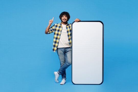 Full body young happy Indian man wear yellow checkered shirt casual clothes big huge blank screen mobile cell phone smartphone with area point index finger up isolated on plain blue cyan background