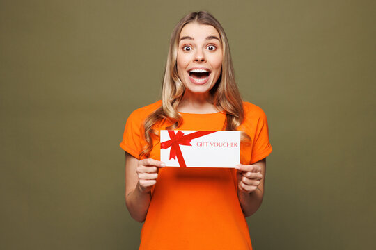 Young shocked surprised woman she wears orange t-shirt casual clothes hold gift certificate coupon voucher card for store isolated on plain pastel green background studio portrait. Lifestyle concept.