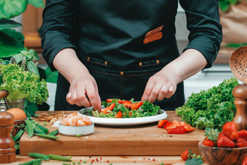Chef carefully assembling vibrant salad with fresh greens, shrimps and strawberries on wooden countertop. Modern cozy kitchen filled with organic produce, creating inviting cooking atmosphere.