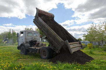 Heavy-duty dump truck is unloading rich soil in a lush green field filled with flowers, under a...