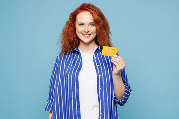 Young smiling happy ginger woman she wear shirt t-shirt casual clothes hold in hand mock up of credit bank card isolated on plain pastel light blue cyan background studio portrait. Lifestyle concept.