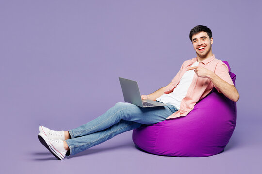 Full body young IT man he wears pink shirt t-shirt casual clothes sit in bag chair hold use work point on laptop pc computer isolated on plain pastel light purple background studio. Lifestyle concept.