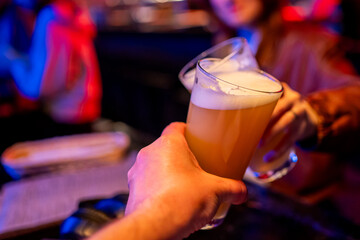 Close-up of two people clinking beer glasses in a lively bar, celebrating and enjoying a cheerful night out with foam-topped drinks in hand.