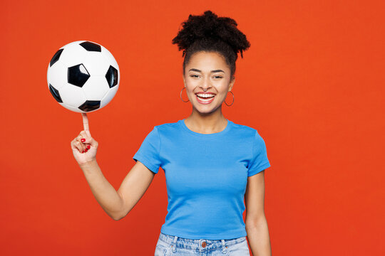 Young smiling happy woman fan she wearing basic blue t-shirt cheer up support football sport team hold in hand spin on finger soccer ball watch tv live stream isolated on plain red orange background.