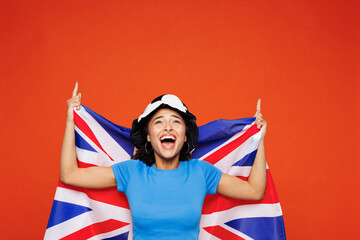 Young woman fan wearing blue t-shirt wrapped in British flag cheer up support football sport team hold soccer ball watch tv live stream point finger overhead isolated on plain red orange background.