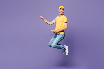 Full body young delivery guy employee man wear yellow cap t-shirt uniform workwear work as dealer courier jump high point index finger aside on area mock up isolated on plain pastel purple background.