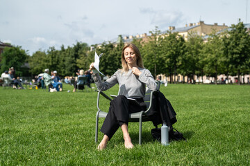 Joyful woman using makeshift paper fan to cool down, sunbathing on grass in hot summer weather. Businesswoman taking break, heat adaptation, solo relax, post work cooling, hydration with water bottle