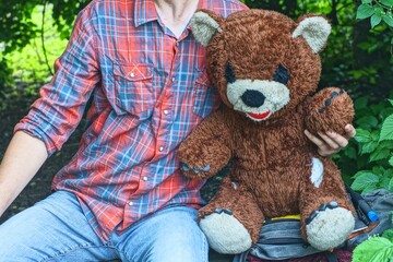  man in checkered shirt and  jeans holding in hand one big old toy soft plush brown baby gift with black eyes and nose and white ears and face torn teddy bear outside