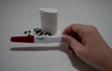 Close-up of a hand holding a pregnancy test with a red and white handle, next to a group of green and white capsules and a white pill container on a plain white background, representing health