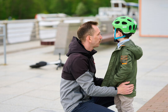 father teaching son safe skateboarding outdoors. man kneeling beside child wearing green helmet, bonding moment captured in urban setting. parental guidance, outdoor activity, family time - Powered by Adobe
