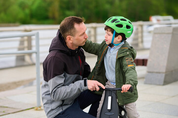 father and son outdoor bonding over scooter. dad kneeling beside young boy, offering encouragement and support. child wearing green helmet, ready for adventure. urban park setting, family time theme