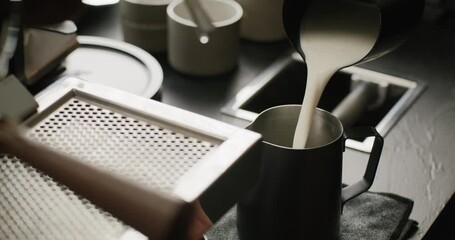 Close-up of milk being poured into a small frothing pitcher, followed by steaming with a steam wand. At the end, the barista gently taps the pitcher on the counter to settle the foam.
