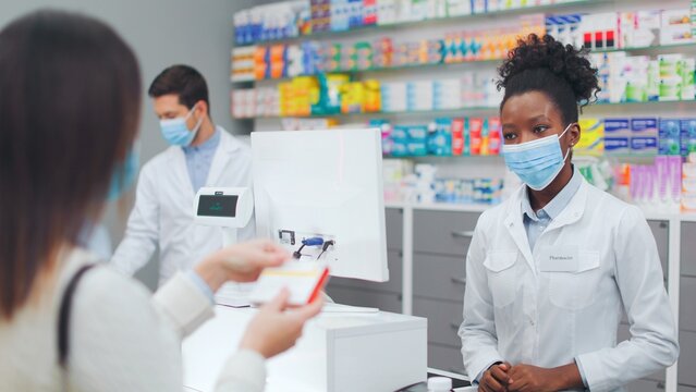 Female client wanting to buy drug by prescription. African American pharmacist picking up medicine for woman. Female druggist wearing protective mask and communicating with client.