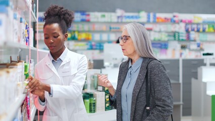 Woman pharmacist advising medicine to female client. African American druggist advising pills to customer. Female pharmacist and customer in pharmacy. Senior woman choosing medication in drugstore.