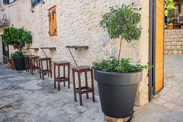 A cozy outdoor bar setup features wooden stools and small tables along a rustic stone wall, adorned with potted trees and greenery, evoking a charming Mediterranean ambiance.