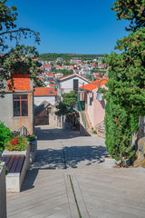 A picturesque view of Primosten, Croatia, showcasing a charming stone street leading down to a vibrant coastal town with traditional red-tiled roofs, framed by lush Mediterranean vegetation