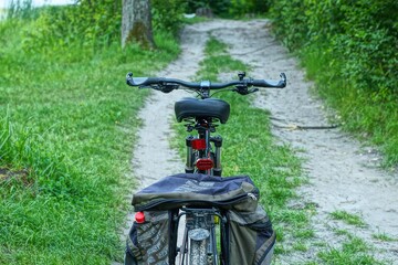 back of black iron sport modern mountain bike with soft saddle and springs handlebar frame rear plastic red reflector and panniers stands on forest road and green grass