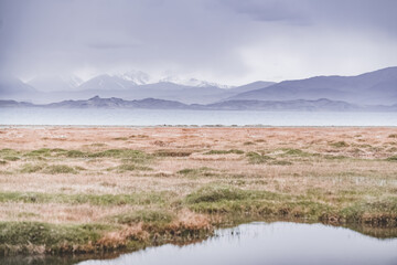 Grass and stone shore of Lake Karakul with blue water and mountain peaks in the background, cloudy...