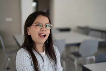 Cute girl in glasses and sweater touches wobbly tooth with tongue while standing in blurred background in school cafeteria. Dental and educational children's concept