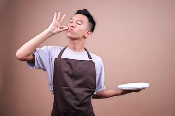 Portrait Of Young Asian Male In Brown Apron Holding Empty White Plate Making Delicious Gesture