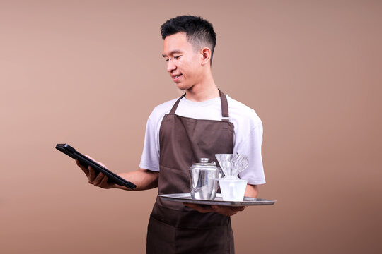 Young Asian Male In Brown Apron Looking At Digital Tablet While Holding Coffee Brewing Set On Tray