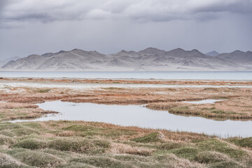 Grass and stone shore of Lake Karakul with blue water and mountain peaks in the background, cloudy weather and sky with clouds and clouds in the Pamirs, panoramic landscape