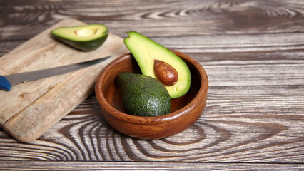 One halved avocado with seed and one whole in a wooden bowl, with a knife and avocado half on a cutting board over rustic wood surface