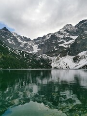 Towering snow-covered Tatra Mountains are reflected in the clear emerald waters of Morskie Oko lake beneath a moody sky near Zakopane, Poland