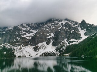 Snow-covered cliffs of the Tatra Mountains tower above Morskie Oko, their reflection mirrored in the lake still waters beneath a cloudy spring sky
