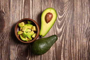 Fresh whole avocado, halved avocado with seed, and cubed pieces in a wooden bowl displayed on a dark rustic wooden background, perfect for food content, top view