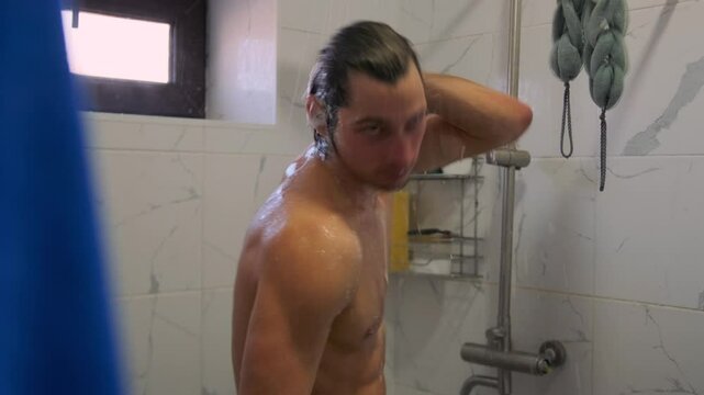A muscular man with wet hair sings in the shower, using the showerhead as a microphone. Dancing with drive and a smile, he turns his routine into an impromptu musical performance.