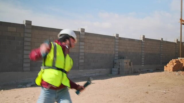A construction worker in a hard hat and safety vest jumps and dances with a drill and saw, lost in the music from his earbuds. A joyful, playful burst of energy and self expression mid workday slacker
