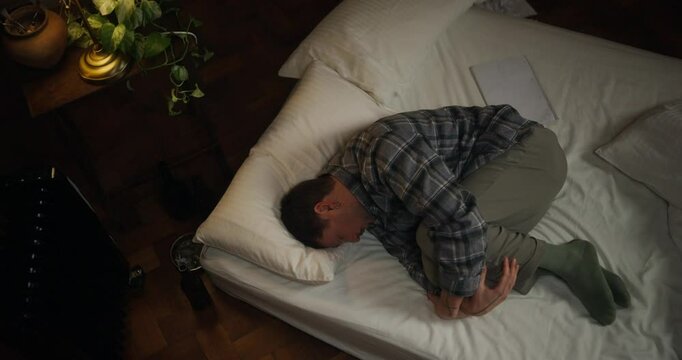 Tired man in checkered shirt lies on mattress and holds knees with hands to rest and let his thoughts wander in evening in cozy room