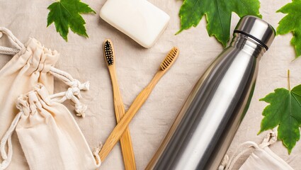 Flat lay of eco-friendly essentials: bamboo toothbrush, stainless steel bottle, cloth bags, and bar soap on neutral background with green leaves.