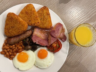 Traditional English breakfast on a white plate on wooden table: 3 hash browns, slices of bacon, baked beans, mushrooms, half a tomato, 2 fried eggs, a Lincolnshire sausage, and a glass of orange juice
