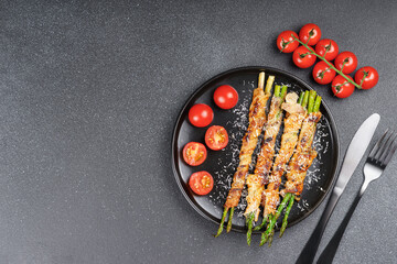 Delicious crispy bacon wrapped asparagus served with fresh cherry tomatoes on black plate and gray background. Perfect food photography for culinary blogs, menus, or healthy meal promotions.