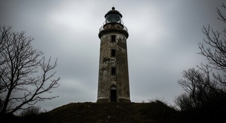 A weathered lighthouse stands tall against a cloudy sky surrounded by bare trees in a desolate landscape