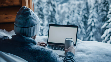 Person in winter attire using a laptop outdoors on a snowy landscape, holding a mug, with a backdrop of snow-covered trees.