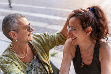 beautiful couple of middle-aged women having fun exchanging tenderness sitting at an outdoor coffee bar in a historic square
