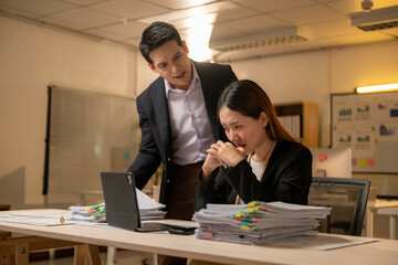A man and a woman are sitting at a desk with a laptop and piles of papers
