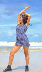 Beautiful Asian woman poses on a beach wearing a stylish floral dress and black ankle boots, set against a backdrop of waves and a partly cloudy sky