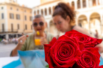 close up of a bunch of red roses in front of a beautiful couple of middle aged female friends enjoying an aperitif sitting outdoors - focus on the roses